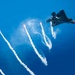 A stealth fighter jet, consistent with the F-22 Raptor design, is captured in flight against a vivid blue sky while deploying multiple flares. The flares trail bright white smoke plumes downward in a dramatic fan pattern. This appears to be an airshow demonstration or a countermeasure deployment exercise.