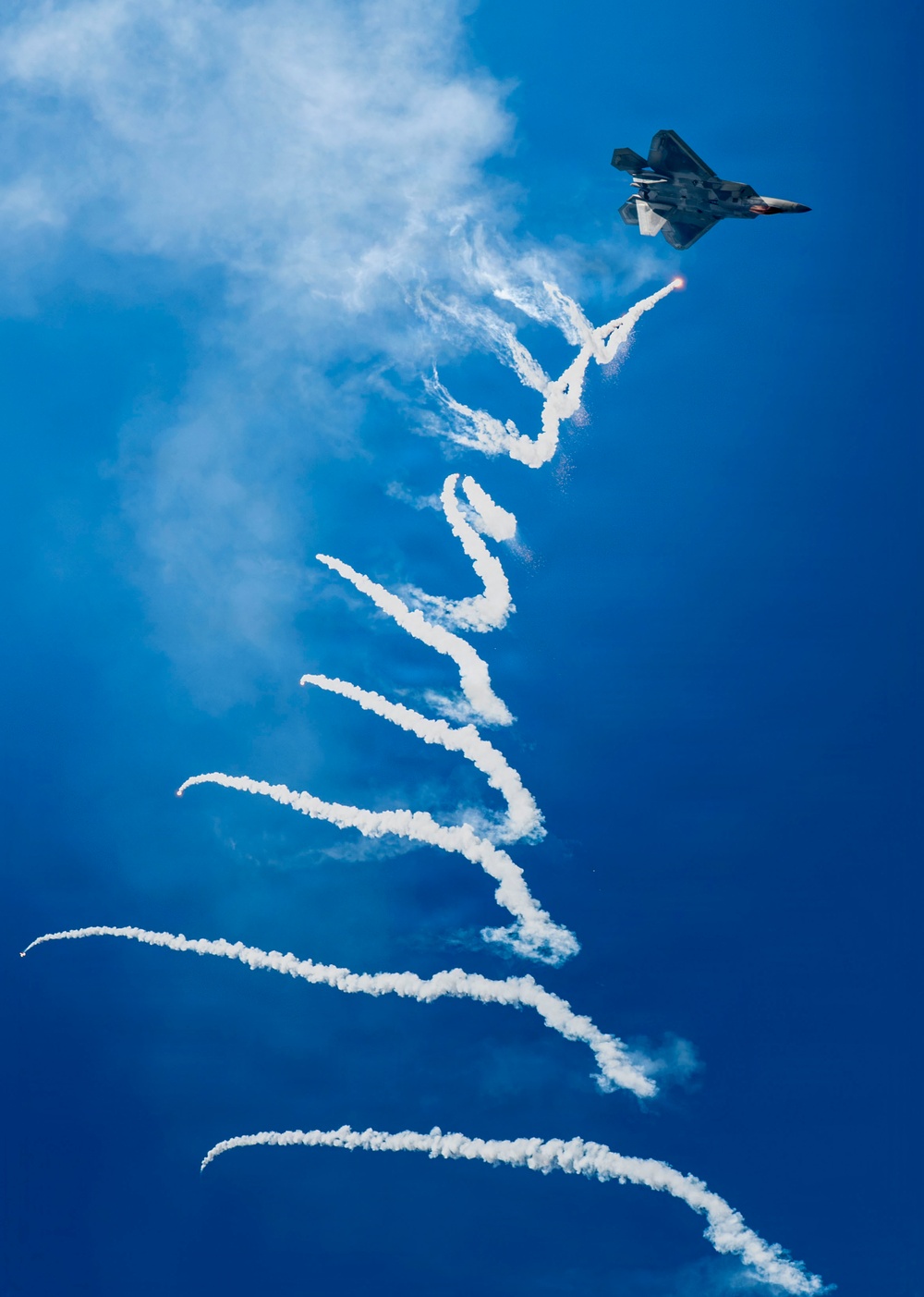 A stealth fighter jet, identifiable as an F-22 Raptor, is captured in flight against a vivid blue sky while dispensing flares and leaving a complex, serpentine smoke trail. The aircraft appears to be performing an aerobatic demonstration, likely at an airshow. The long, winding white smoke trail extends dramatically downward through most of the frame, creating a visually striking pattern.