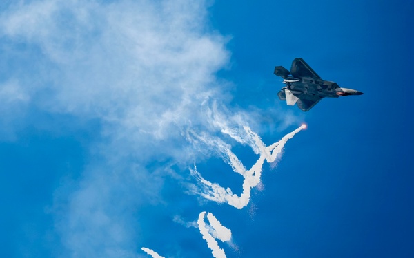 A stealth fighter jet, identifiable as an F-22 Raptor, is captured in flight against a vivid blue sky while dispensing flares and leaving a complex, serpentine smoke trail. The aircraft appears to be performing an aerobatic demonstration, likely at an airshow. The long, winding white smoke trail extends dramatically downward through most of the frame, creating a visually striking pattern.