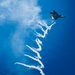 A stealth fighter jet, identifiable as an F-22 Raptor, is captured in flight against a vivid blue sky while dispensing flares and leaving a complex, serpentine smoke trail. The aircraft appears to be performing an aerobatic demonstration, likely at an airshow. The long, winding white smoke trail extends dramatically downward through most of the frame, creating a visually striking pattern.