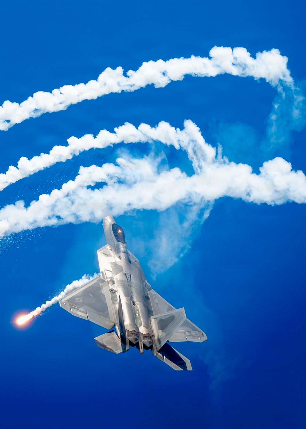 A fifth-generation stealth fighter jet, consistent with an F-22 Raptor, is captured in a steep climbing maneuver against a vivid blue sky. The aircraft is actively deploying flares from its left side, producing bright orange flames and white smoke trails. The smoke trails from previous maneuvers form sweeping, looping patterns across the upper portion of the image.