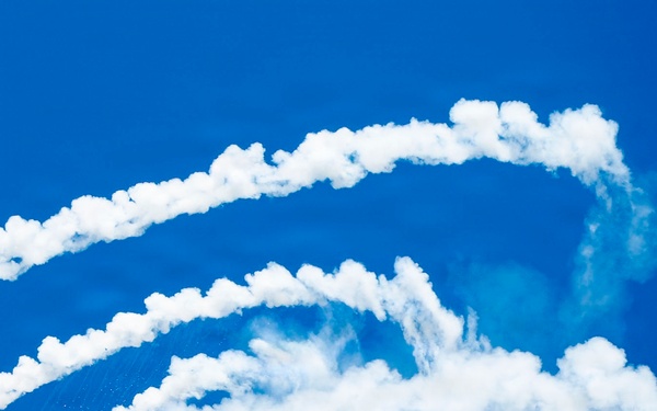 A fifth-generation stealth fighter jet, consistent with an F-22 Raptor, is captured in a steep climbing maneuver against a vivid blue sky. The aircraft is actively deploying flares from its left side, producing bright orange flames and white smoke trails. The smoke trails from previous maneuvers form sweeping, looping patterns across the upper portion of the image.