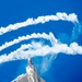 A fifth-generation stealth fighter jet, consistent with an F-22 Raptor, is captured in a steep climbing maneuver against a vivid blue sky. The aircraft is actively deploying flares from its left side, producing bright orange flames and white smoke trails. The smoke trails from previous maneuvers form sweeping, looping patterns across the upper portion of the image.