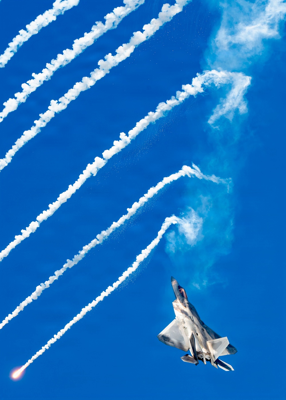 A fifth-generation stealth fighter jet, identifiable as an F-22 Raptor, is captured in a steep banking maneuver against a vivid blue sky. The aircraft is dispensing multiple flares, which leave bright white smoke trails arcing diagonally across the frame. One flare near the bottom-left corner is still visibly burning with an orange flame, suggesting this is an airshow demonstration.