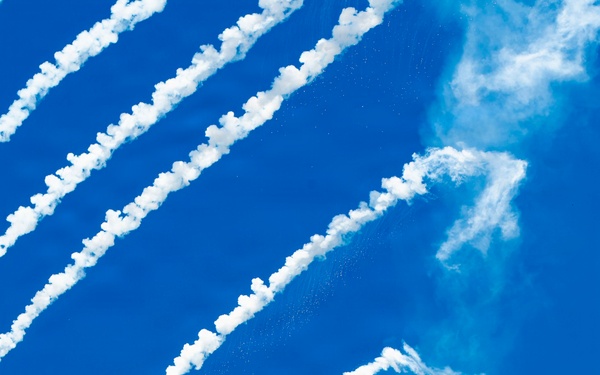 A fifth-generation stealth fighter jet, identifiable as an F-22 Raptor, is captured in a steep banking maneuver against a vivid blue sky. The aircraft is dispensing multiple flares, which leave bright white smoke trails arcing diagonally across the frame. One flare near the bottom-left corner is still visibly burning with an orange flame, suggesting this is an airshow demonstration.