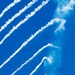 A fifth-generation stealth fighter jet, identifiable as an F-22 Raptor, is captured in a steep banking maneuver against a vivid blue sky. The aircraft is dispensing multiple flares, which leave bright white smoke trails arcing diagonally across the frame. One flare near the bottom-left corner is still visibly burning with an orange flame, suggesting this is an airshow demonstration.