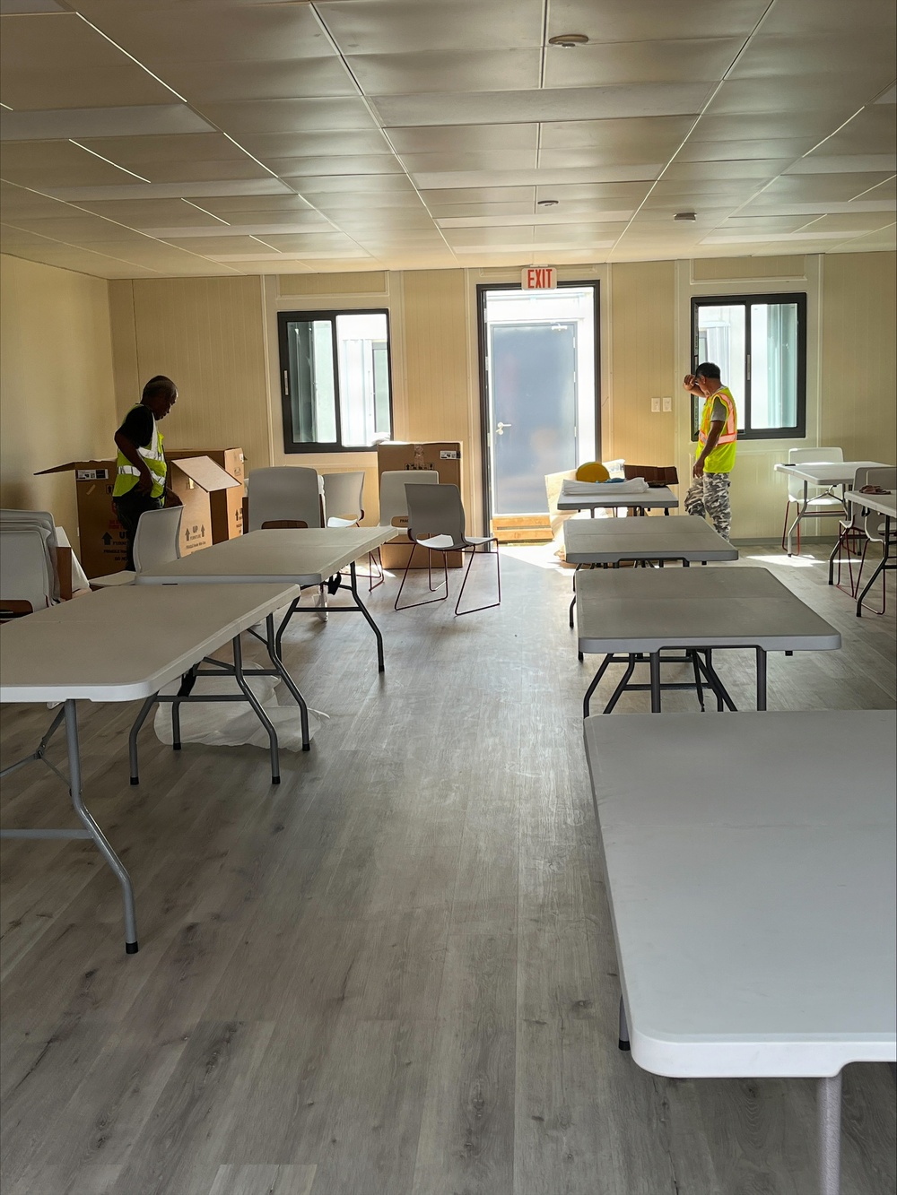 The image shows the interior of a modular or prefabricated building being set up, likely as a temporary facility such as a community center, shelter, or dining hall. Two workers wearing high-visibility safety vests are actively arranging furniture and unpacking cardboard boxes. Multiple white folding tables and chairs are being positioned throughout the room, which features laminate flooring, panel ceiling tiles, and an illuminated EXIT sign above the central door.