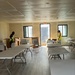The image shows the interior of a modular or prefabricated building being set up, likely as a temporary facility such as a community center, shelter, or dining hall. Two workers wearing high-visibility safety vests are actively arranging furniture and unpacking cardboard boxes. Multiple white folding tables and chairs are being positioned throughout the room, which features laminate flooring, panel ceiling tiles, and an illuminated EXIT sign above the central door.