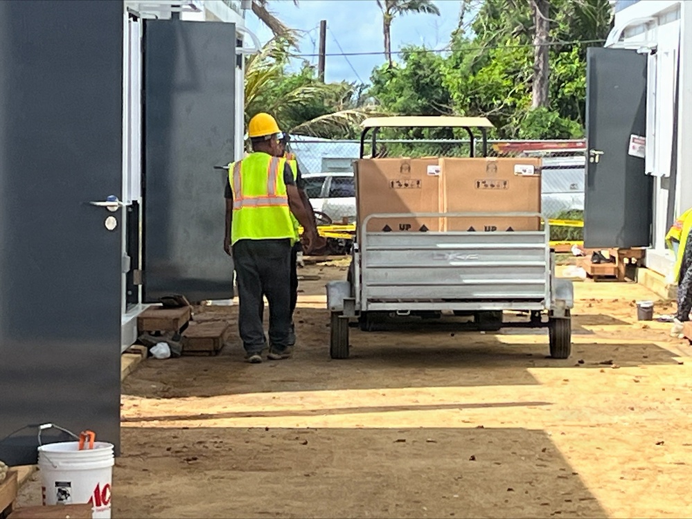 A construction worker wearing a yellow hard hat and high-visibility vest is walking alongside a motorized cart loaded with large cardboard boxes on a dirt construction site. The site appears to be located in a tropical environment, with palm trees and lush vegetation visible in the background. The area shows signs of active construction or post-storm recovery work, with debris and building materials scattered around.