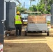 A construction worker wearing a yellow hard hat and high-visibility vest is walking alongside a motorized cart loaded with large cardboard boxes on a dirt construction site. The site appears to be located in a tropical environment, with palm trees and lush vegetation visible in the background. The area shows signs of active construction or post-storm recovery work, with debris and building materials scattered around.