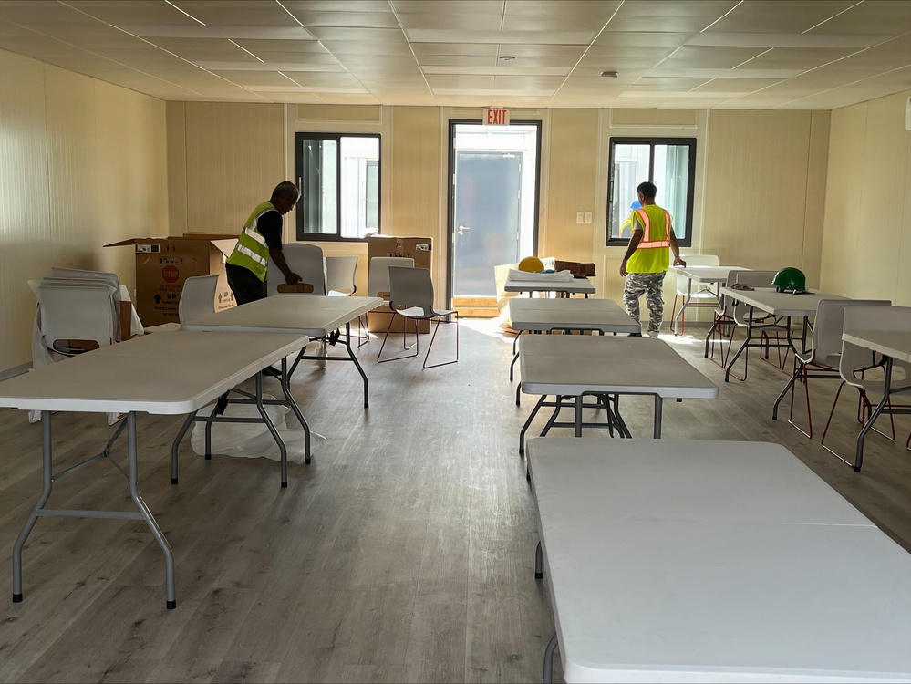 Two workers in high-visibility safety vests are setting up a room with folding tables and chairs, likely in a modular or prefabricated building. The space appears to be a break room, cafeteria, or meeting area under preparation. Cardboard boxes and hard hats are visible, suggesting the setup is still in progress.