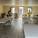 Two workers in high-visibility safety vests are setting up a room with folding tables and chairs, likely in a modular or prefabricated building. The space appears to be a break room, cafeteria, or meeting area under preparation. Cardboard boxes and hard hats are visible, suggesting the setup is still in progress.