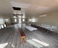 The image shows a multipurpose interior room with folding tables and chairs arranged along the walls and in the center, suggesting a meeting or event space. The room features a drop ceiling with several missing or damaged tiles, a wall-mounted mini-split air conditioning unit, and new-looking laminate flooring. The space appears to be in a transitional or partially renovated state.