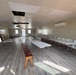 The image shows a multipurpose interior room with folding tables and chairs arranged along the walls and in the center, suggesting a meeting or event space. The room features a drop ceiling with several missing or damaged tiles, a wall-mounted mini-split air conditioning unit, and new-looking laminate flooring. The space appears to be in a transitional or partially renovated state.