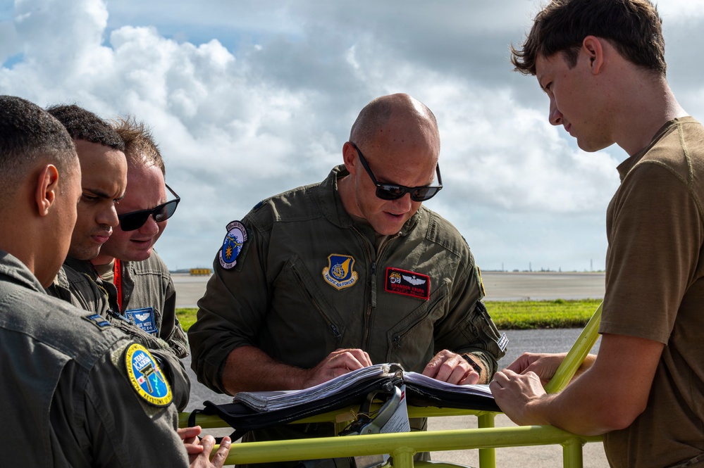 A group of military personnel, appearing to be U.S. Air Force aviators, are gathered around a yellow cart or stand reviewing documents or a flight binder outdoors on what appears to be an airfield. The central figure is a bald man wearing a green flight suit with Pacific Air Forces patches and pilot wings. The setting suggests a pre-flight briefing or mission planning activity on a flight line.
