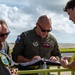 A group of military personnel, appearing to be U.S. Air Force aviators, are gathered around a yellow cart or stand reviewing documents or a flight binder outdoors on what appears to be an airfield. The central figure is a bald man wearing a green flight suit with Pacific Air Forces patches and pilot wings. The setting suggests a pre-flight briefing or mission planning activity on a flight line.