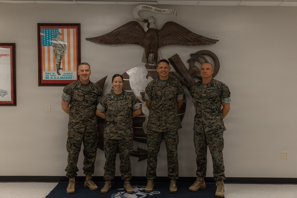 Four U.S. Marines in woodland MARPAT digital camouflage uniforms pose together in front of a large Eagle, Globe, and Anchor (EGA) emblem mounted on a wall. The setting appears to be an official Marine Corps facility, likely a recruiting station or administrative building. The group consists of three males and one female, all standing at ease and smiling.