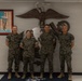 Four U.S. Marines in woodland MARPAT digital camouflage uniforms pose together in front of a large Eagle, Globe, and Anchor (EGA) emblem mounted on a wall. The setting appears to be an official Marine Corps facility, likely a recruiting station or administrative building. The group consists of three males and one female, all standing at ease and smiling.