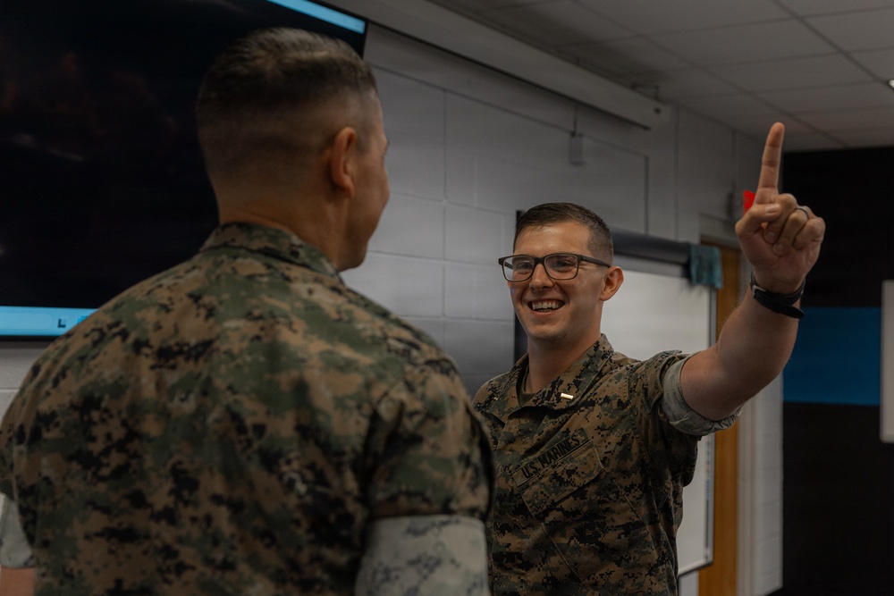 Two U.S. Marines in MARPAT camouflage uniforms are engaged in a conversation inside what appears to be a briefing or classroom environment. The Marine facing the camera is smiling broadly and holding up one finger, possibly making a point or emphasizing something during the discussion. The setting suggests a training or operational planning context.