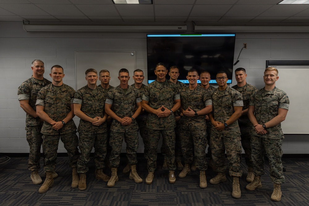A group of approximately 13 military personnel in U.S. Marine Corps MARPAT camouflage uniforms pose for a formal group photograph indoors. They are standing in a single row in what appears to be a briefing room or classroom facility. The group appears to be posing after a meeting or training event.