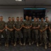 A group of approximately 13 military personnel in U.S. Marine Corps MARPAT camouflage uniforms pose for a formal group photograph indoors. They are standing in a single row in what appears to be a briefing room or classroom facility. The group appears to be posing after a meeting or training event.