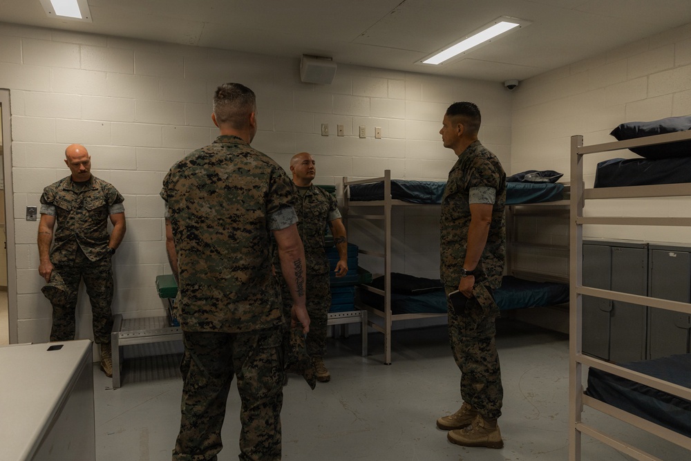 Four military personnel in MARPAT (Marine Corps digital camouflage) uniforms are gathered inside what appears to be a military barracks room. They appear to be engaged in a briefing, inspection, or discussion. The room contains metal bunk beds with dark bedding and storage shelving units along the walls.