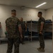 Four military personnel in MARPAT (Marine Corps digital camouflage) uniforms are gathered inside what appears to be a military barracks room. They appear to be engaged in a briefing, inspection, or discussion. The room contains metal bunk beds with dark bedding and storage shelving units along the walls.