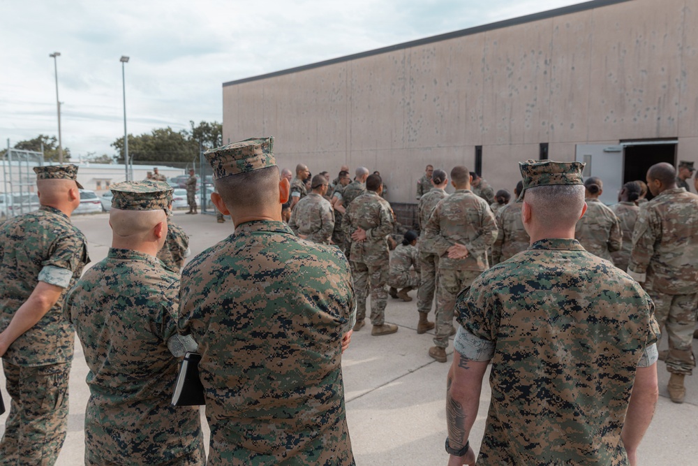 A group of military personnel in two distinct camouflage uniforms are gathered outdoors near a large concrete building. The foreground shows individuals in USMC MARPAT (Marine Pattern) digital camouflage, while the larger group in the background appears to wear OCP (Operational Camouflage Pattern) uniforms typical of Army or Air Force personnel. The gathering appears to be a briefing, formation, or joint training event.