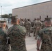 A group of military personnel in two distinct camouflage uniforms are gathered outdoors near a large concrete building. The foreground shows individuals in USMC MARPAT (Marine Pattern) digital camouflage, while the larger group in the background appears to wear OCP (Operational Camouflage Pattern) uniforms typical of Army or Air Force personnel. The gathering appears to be a briefing, formation, or joint training event.