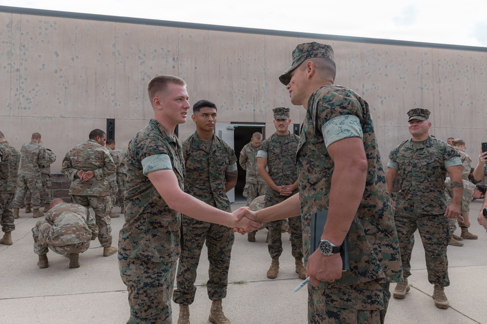 A group of military personnel in camouflage uniforms are gathered outdoors on a paved area near a concrete building. Two individuals in the foreground are shaking hands, suggesting a formal recognition or greeting ceremony. The group includes both U.S. Marine Corps personnel (in MARPAT digital camouflage) and what appear to be U.S. Army soldiers (in OCP/MultiCam pattern) in the background.