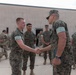 A group of military personnel in camouflage uniforms are gathered outdoors on a paved area near a concrete building. Two individuals in the foreground are shaking hands, suggesting a formal recognition or greeting ceremony. The group includes both U.S. Marine Corps personnel (in MARPAT digital camouflage) and what appear to be U.S. Army soldiers (in OCP/MultiCam pattern) in the background.