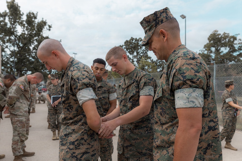 A group of U.S. military personnel in digital camouflage uniforms are gathered outdoors with heads bowed, appearing to be engaged in a prayer or moment of reflection. The foreground shows Marines in MARPAT (Marine Pattern) uniforms, while personnel in Army ACU-pattern uniforms are visible in the background. The setting appears to be a military installation or base.