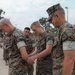 A group of U.S. military personnel in digital camouflage uniforms are gathered outdoors with heads bowed, appearing to be engaged in a prayer or moment of reflection. The foreground shows Marines in MARPAT (Marine Pattern) uniforms, while personnel in Army ACU-pattern uniforms are visible in the background. The setting appears to be a military installation or base.
