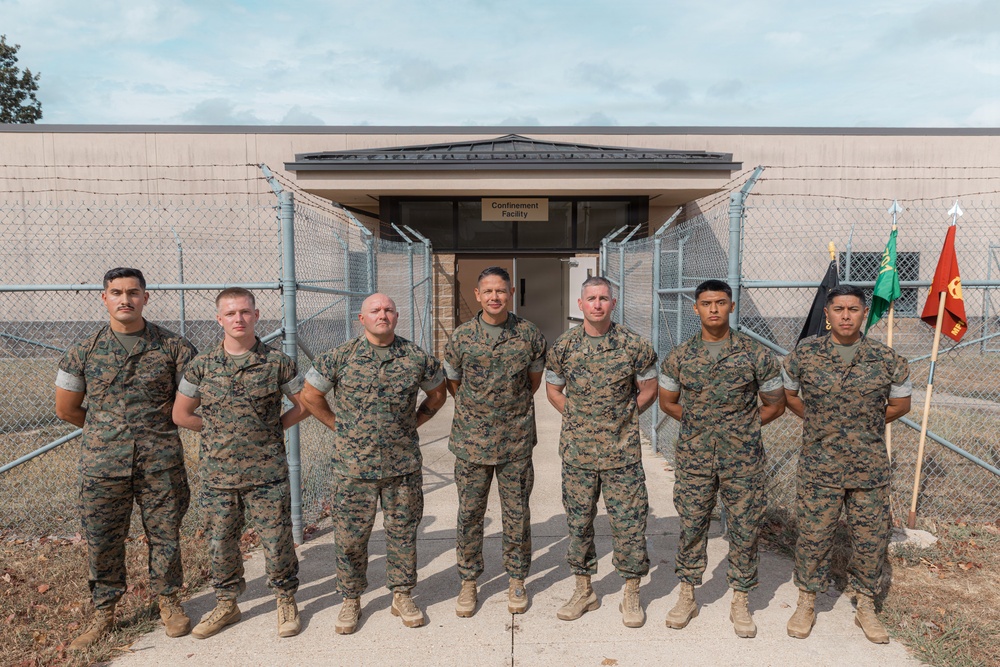 Seven U.S. Marines in woodland MARPAT camouflage uniforms pose for a group photo in front of a building labeled 'Confinement Facility.' They are standing on a concrete walkway flanked by chain-link fencing with razor wire. Several military unit guidon flags are visible on the right side of the group.