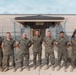 Seven U.S. Marines in woodland MARPAT camouflage uniforms pose for a group photo in front of a building labeled 'Confinement Facility.' They are standing on a concrete walkway flanked by chain-link fencing with razor wire. Several military unit guidon flags are visible on the right side of the group.