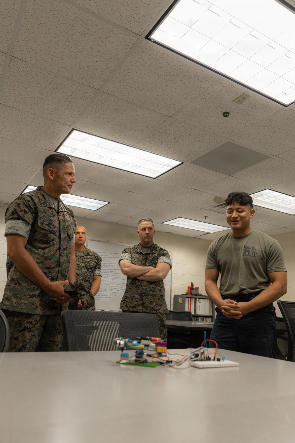 A group of U.S. Marines in camouflage uniforms are gathered around a table with a young man in a military physical training (PT) shirt. The young man appears to be presenting or demonstrating a project involving LEGO components and an electronics breadboard with wiring. The setting is an indoor classroom or conference room environment.