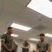 A group of U.S. Marines in camouflage uniforms are gathered around a table with a young man in a military physical training (PT) shirt. The young man appears to be presenting or demonstrating a project involving LEGO components and an electronics breadboard with wiring. The setting is an indoor classroom or conference room environment.