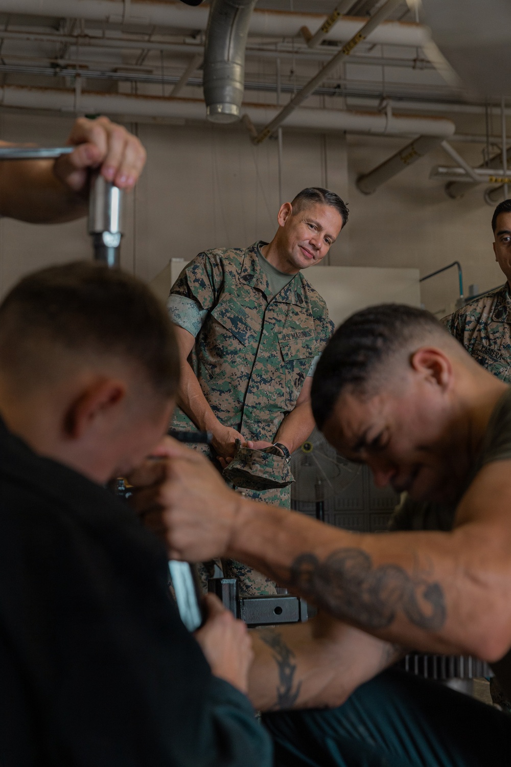 A U.S. Marine in MARPAT camouflage uniform observes personnel working in what appears to be a maintenance or technical workspace. Two individuals in the foreground, one shirtless with visible tattoos, appear to be working with tools or equipment. A second uniformed Marine is partially visible in the background.