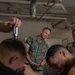 A U.S. Marine in MARPAT camouflage uniform observes personnel working in what appears to be a maintenance or technical workspace. Two individuals in the foreground, one shirtless with visible tattoos, appear to be working with tools or equipment. A second uniformed Marine is partially visible in the background.