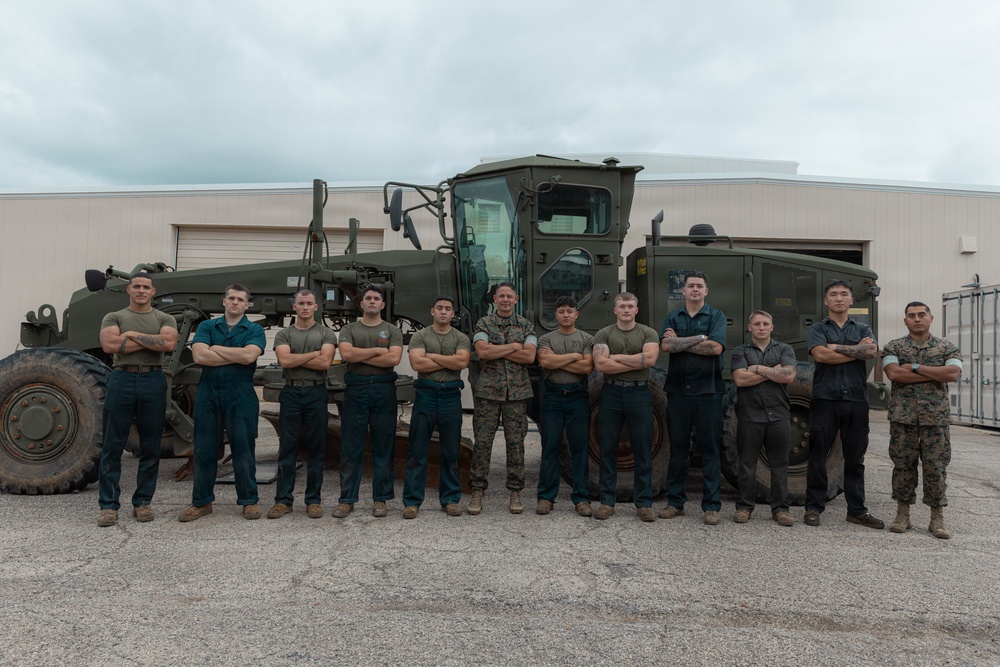 A group of approximately 13 military personnel, appearing to be U.S. Marines based on uniforms, pose in a group photo with arms crossed in front of a large olive-drab military motor grader or road grading vehicle. The photo appears to be a formal unit or team photograph taken outdoors on a paved surface near a military facility building. Personnel are wearing a mix of camouflage utilities and dark blue/green work coveralls.