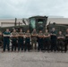 A group of approximately 13 military personnel, appearing to be U.S. Marines based on uniforms, pose in a group photo with arms crossed in front of a large olive-drab military motor grader or road grading vehicle. The photo appears to be a formal unit or team photograph taken outdoors on a paved surface near a military facility building. Personnel are wearing a mix of camouflage utilities and dark blue/green work coveralls.