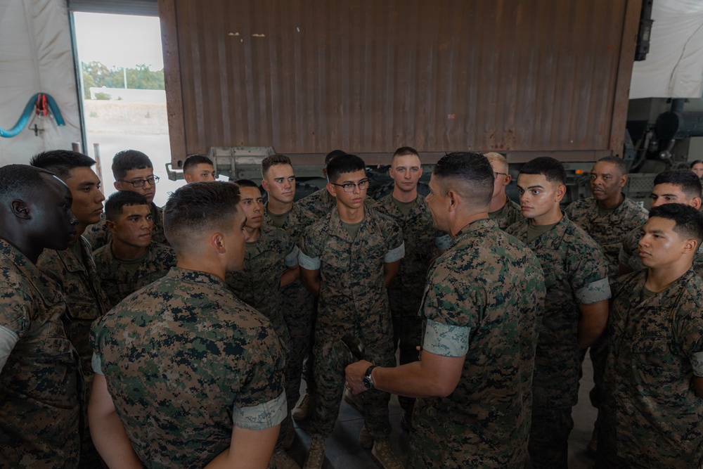 A group of approximately 20 military personnel in U.S. Marine Corps MARPAT (Marine Pattern) camouflage uniforms are gathered in what appears to be a briefing or huddle inside a hangar or warehouse facility. One individual, facing away from the camera, appears to be addressing the group, suggesting a leadership briefing or pre-mission instruction. The setting appears to be an operational or training environment.