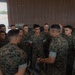 A group of approximately 20 military personnel in U.S. Marine Corps MARPAT (Marine Pattern) camouflage uniforms are gathered in what appears to be a briefing or huddle inside a hangar or warehouse facility. One individual, facing away from the camera, appears to be addressing the group, suggesting a leadership briefing or pre-mission instruction. The setting appears to be an operational or training environment.