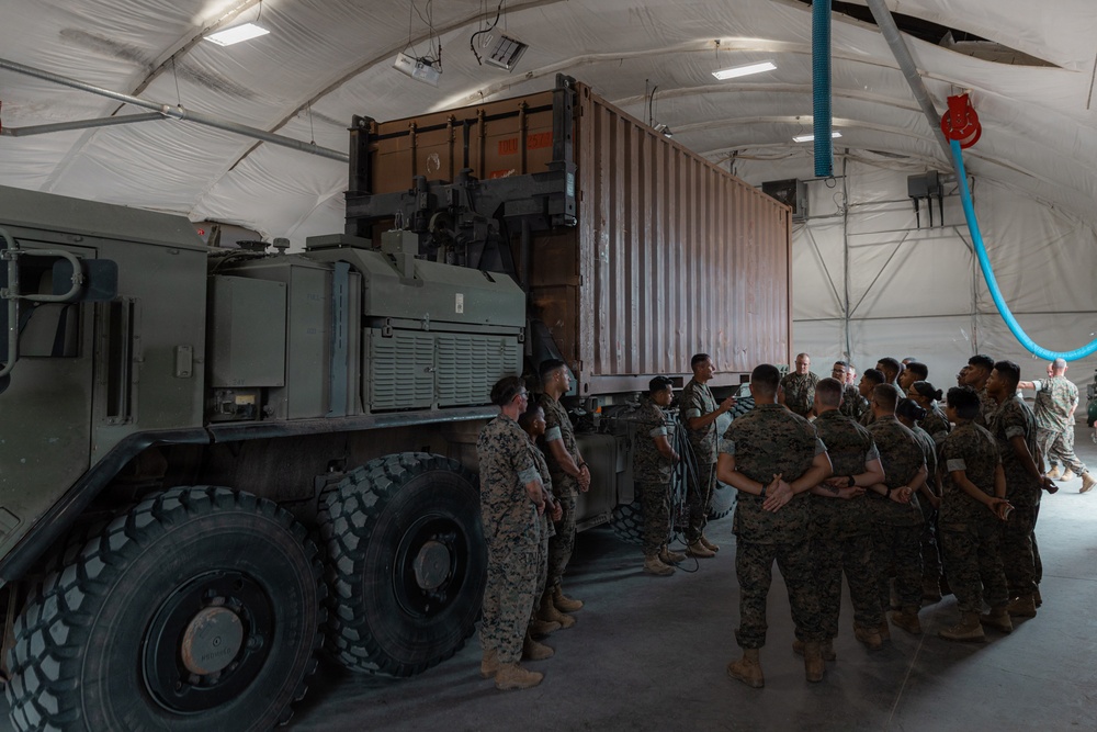A group of military personnel in camouflage uniforms are gathered inside a large fabric-covered hangar or temporary structure. They appear to be receiving a briefing or instruction near a large military heavy-duty truck carrying a shipping container. The setting suggests a logistics or equipment training exercise.