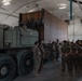A group of military personnel in camouflage uniforms are gathered inside a large fabric-covered hangar or temporary structure. They appear to be receiving a briefing or instruction near a large military heavy-duty truck carrying a shipping container. The setting suggests a logistics or equipment training exercise.