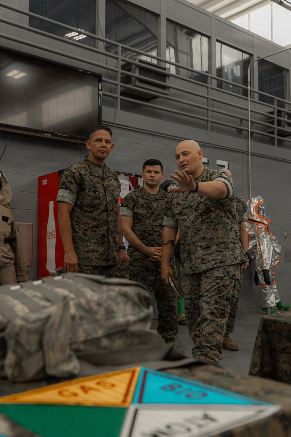 Three military personnel in MARPAT (Marine digital camouflage) uniforms are gathered in an indoor facility, with one individual actively gesturing and appearing to brief or instruct the others. The setting includes hazmat/proximity suits visible in the background, warning placards on a table in the foreground, and a Coca-Cola vending machine against the wall. The scene suggests a CBRN (Chemical, Biological, Radiological, Nuclear) or hazardous materials training briefing.