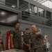 Three military personnel in MARPAT (Marine digital camouflage) uniforms are gathered in an indoor facility, with one individual actively gesturing and appearing to brief or instruct the others. The setting includes hazmat/proximity suits visible in the background, warning placards on a table in the foreground, and a Coca-Cola vending machine against the wall. The scene suggests a CBRN (Chemical, Biological, Radiological, Nuclear) or hazardous materials training briefing.
