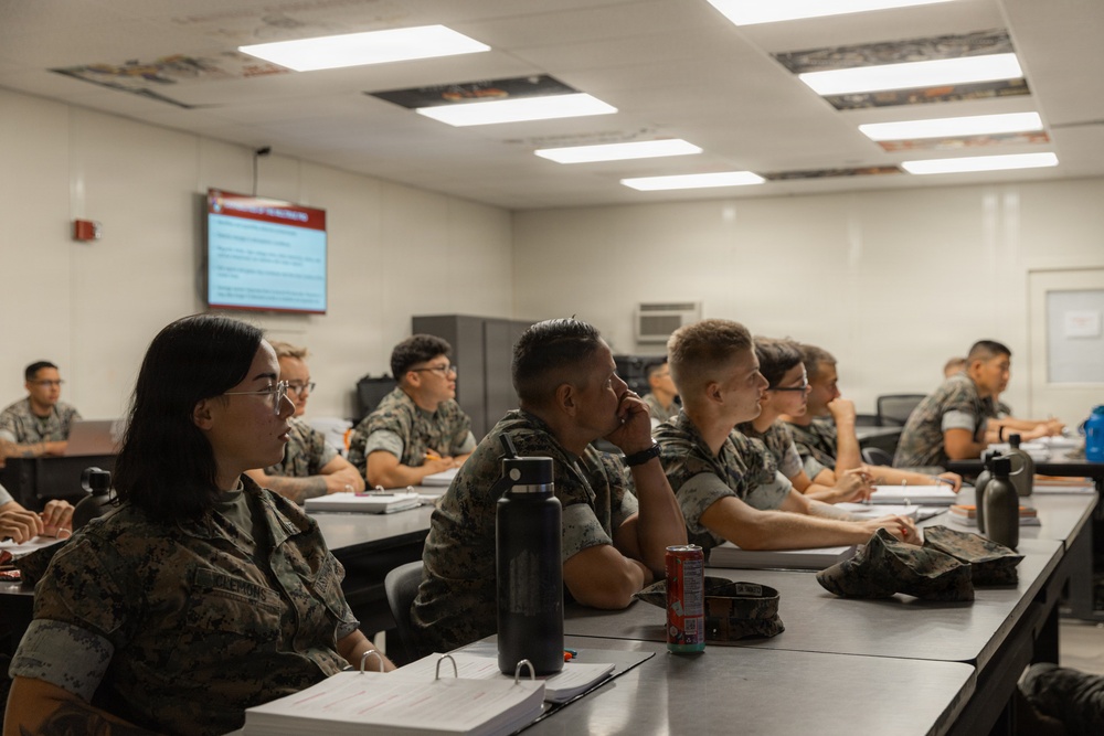 A group of military personnel in camouflage uniforms are seated at desks in a classroom setting, attentively watching a presentation on a wall-mounted screen. The setting appears to be a formal military training or educational environment. Multiple students are visible in rows, with notebooks, water bottles, and personal items on their desks.