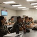 A group of military personnel in camouflage uniforms are seated at desks in a classroom setting, attentively watching a presentation on a wall-mounted screen. The setting appears to be a formal military training or educational environment. Multiple students are visible in rows, with notebooks, water bottles, and personal items on their desks.