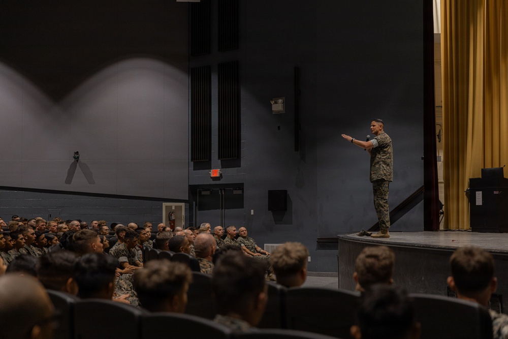 A military officer in camouflage uniform stands on a stage addressing a large audience of uniformed military personnel seated in an auditorium. The speaker is gesturing with one arm extended, appearing to be delivering a briefing or command address. The setting is a formal military theater or auditorium with stage lighting and gold curtains.